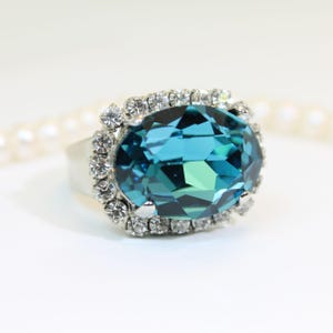 May include: A silver-toned ring featuring a large, oval-cut teal gemstone as the centerpiece. The central stone is surrounded by a halo of small, clear, round-cut white crystals set in a metallic frame, creating a bright, reflective appearance.