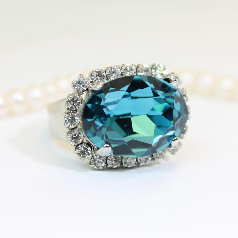 May include: A silver-toned ring featuring a large, oval-cut teal gemstone as the centerpiece. The central stone is surrounded by a halo of small, clear, round-cut white crystals set in a metallic frame, creating a bright, reflective appearance.