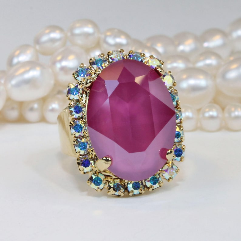 May include: A large, oval-shaped, pink gemstone ring with a gold setting. The ring is surrounded by a halo of smaller, iridescent blue gemstones.
