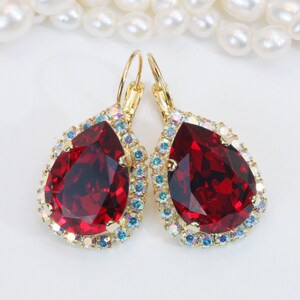 May include: A pair of gold dangle earrings with a large teardrop-shaped red crystal surrounded by smaller clear crystals.