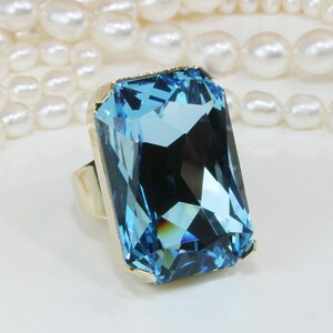May include: A large, rectangular, light blue gemstone ring with a gold-colored band. The gemstone is faceted, reflecting light. The ring is set against a backdrop of white pearls.
