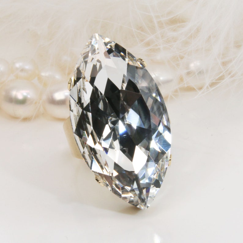May include: A large, clear crystal ring with a marquise cut. The ring is set in a gold band and is surrounded by a white background.