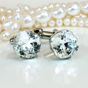May include: A pair of silver-toned cufflinks featuring large, clear, faceted cushion-cut crystals. The cufflinks are positioned in front of a strand of white pearls, showcasing a reflective metallic finish and brilliant light-catching surfaces.