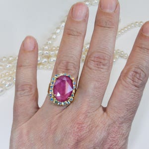 May include: A gold ring with a large, oval-shaped pink gemstone surrounded by a halo of smaller blue gemstones.