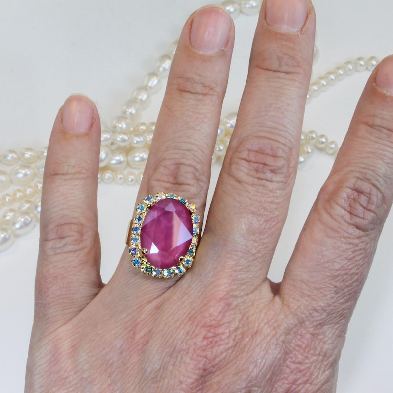 May include: A gold ring with a large, oval-shaped pink gemstone surrounded by a halo of smaller blue gemstones.