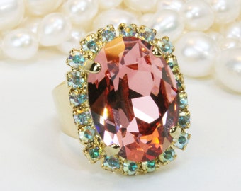 May include: A gold ring with a large oval-shaped pink gemstone surrounded by a halo of smaller clear and green gemstones.