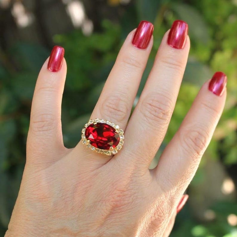May include: A gold ring featuring a large, oval-cut, deep red gemstone as the centerpiece. The central red stone is surrounded by a halo of small, clear, sparkling stones set into the gold band, creating a bright and reflective appearance.