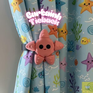 Puede incluir: Un recogedor de cortina con forma de estrella de mar rosa está sujeto a una cortina azul con un patrón repetido de estrellas de mar y vida marina. El recogedor está hecho de un material impreso en 3D y tiene una cara sonriente. El texto "Curtain Tieback" es rosa neón.
