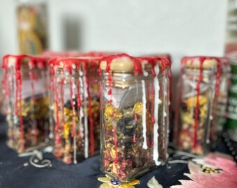 Op de afbeelding: Een groep kleine glazen potten gevuld met gedroogde kruiden en bloemen. De potten zijn bedekt met rode was die aan de zijkanten is gedruppeld.