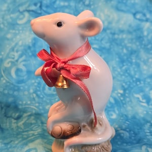 May include: A ceramic figurine of a mouse in a standing pose. The mouse is light pink with a red ribbon tied around its neck, and a small gold bell. The mouse is perched on a brown base.