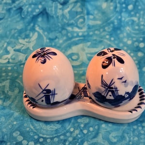 May include: A pair of egg-shaped ceramic salt and pepper shakers featuring hand-painted blue windmill scenes on a white background. The shakers sit on a matching white ceramic base with blue decorative trim, and each shaker has a floral pattern on top.