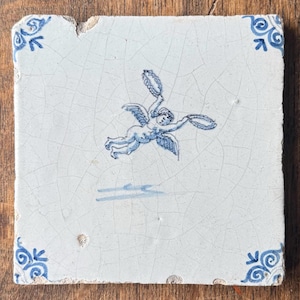Op de afbeelding: Een vierkante keramische tegel met een witte basis en een blauwe craquelé glazuurafwerking. Het midden toont een blauwe illustratie van een gevleugelde cherubijn die een krans vasthoudt. Elke hoek heeft een blauw decoratief krulmotief.