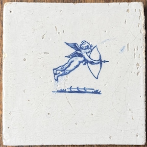 May include: Square white tile with a blue illustration of Cupid, a winged cherub, aiming a bow and arrow. The tile has a slightly distressed appearance, with visible imperfections and a rough edge.