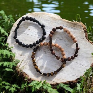 May include: Three beaded bracelets on a wooden surface. The bracelets are made of black lava rock, brown wood, and tiger's eye.