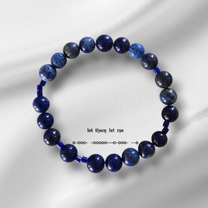 Puede incluir: Un brazalete de cuentas de lapislázuli azul con cuentas espaciadoras azul oscuro. El brazalete está sobre un fondo blanco.
