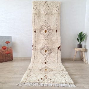 Op de afbeelding: Crèmekleurig, rechthoekig tapijt met een ruitpatroon in bruin en goud. Het tapijt heeft een shaggy textuur en franjes aan de onderkant. Het tapijt wordt gepresenteerd tegen een witte muur.