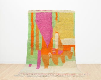 May include: A colorful, abstract rug with a cream fringe. The rug features a vibrant design with pink, orange, green, and yellow geometric shapes. The rug is made of a thick, textured material.