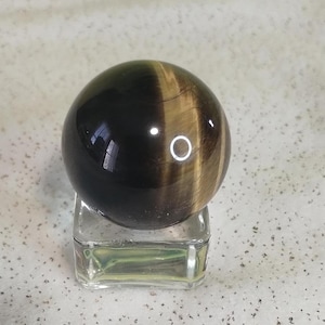 May include: A polished tiger's eye sphere, resting on a clear glass stand. The gemstone is a mix of dark brown and golden-yellow hues, with a glossy finish. The sphere is a decorative item.