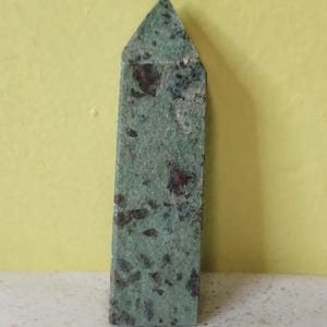 May include: A green and red ruby zoisite crystal obelisk. The stone has a rectangular shape with a pointed top. The surface is textured with dark green and red inclusions. The obelisk is displayed on a white surface against a yellow background.