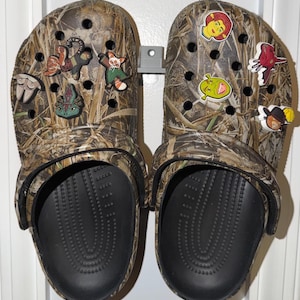 May include: Pair of camouflage Crocs decorated with various cartoon character charms. The Crocs have black soles and are mounted on a gray holder. The charms include Shrek, a red-haired woman, and other colorful figures.