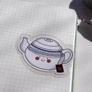 May include: A white ceramic teapot sticker with a blue and white patterned design. The teapot has a smiling face and a red heart on the tea bag.
