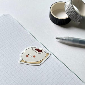 May include: A sticker of a cute, cartoon-style bao bun with a red bean filling, sitting on a yellow square. The sticker is on a grid-lined paper, with black and white patterned washi tape and a pen in the background.
