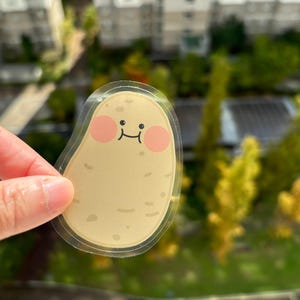 May include: A clear sticker with a cartoon potato design. The potato has a smiling face with pink cheeks and a brown outline. The sticker is on a blurry background of green trees and buildings.