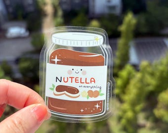 May include: Clear sticker featuring a jar of Nutella with a white lid. The jar is filled with brown Nutella and has a white label with the word "NUTELLA" and the phrase "on everything". The sticker has a cute design with a smiling face and illustrations of a hazelnut and a bread slice.