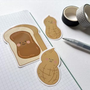 May include: Three cute cartoon stickers featuring a slice of bread with peanut butter, and two peanuts. The peanut stickers have smiling faces and the peanut butter is spread on the bread.