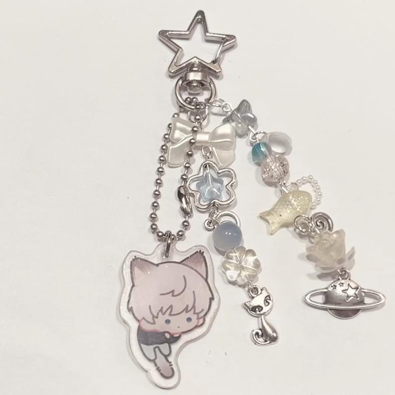 Peut inclure: Un porte-cl&eacute;s avec un charme en argent repr&eacute;sentant un personnage de dessin anim&eacute;, un n&oelig;ud blanc, une &eacute;toile bleue, un charme de chat argent&eacute;, un charme de plan&egrave;te argent&eacute; et d'autres charmes argent&eacute;s et bleus.