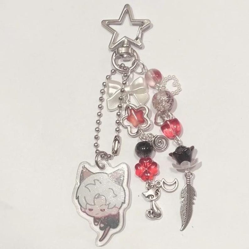 Peut inclure: Un porte-cl&eacute;s en forme d'&eacute;toile argent&eacute;e avec une vari&eacute;t&eacute; de breloques, dont un personnage de dessin anim&eacute; rouge et blanc, un n&oelig;ud blanc, des perles rouges et argent&eacute;es et une plume argent&eacute;e.