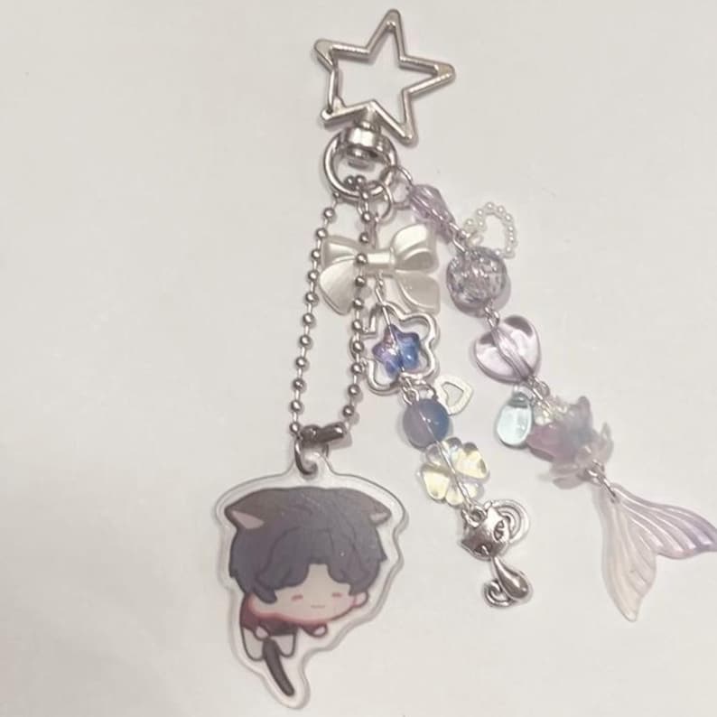 Peut inclure: Un porte-cl&eacute;s en argent avec un charme de personnage de dessin anim&eacute;, un charme d'&eacute;toile, un charme de n&oelig;ud et plusieurs autres charmes. Les charmes sont en verre et en m&eacute;tal et sont dans des tons de violet, bleu et blanc.