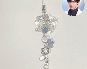 May include: A clear, floral-shaped charm with blue and white accents, featuring a string for attachment. The charm has a delicate design with a snowflake and a teardrop-shaped crystal at the bottom. The text "douhwuadays" is visible.