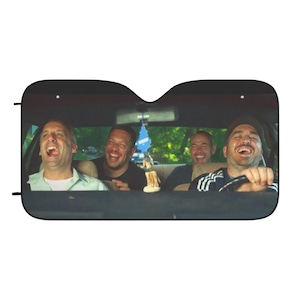 May include: Car sunshade featuring a photograph of four men laughing inside a car. The driver is wearing a black cap and holding the steering wheel. The other three men are in the passenger seats. A blue air freshener hangs from the rearview mirror.