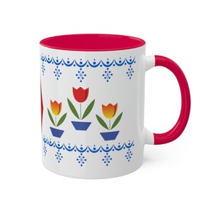 May include: White ceramic mug with a red rim and handle. The mug features a design of three tulips in blue pots with a blue and white snowflake pattern around the top.