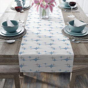 May include: A white table runner featuring a repeating pattern of blue and grey starbursts. The runner is laid on a wooden table set for a meal, with plates, bowls, cutlery, and wine glasses. A vase of pink lilies is in the centre.