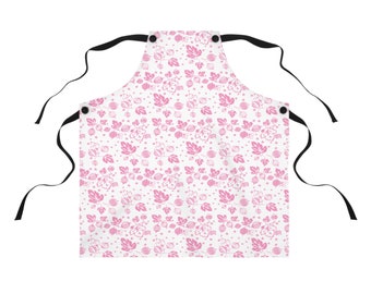 May include: White apron with a pink floral pattern. The apron features a repeating design of leaves, berries, and dots. It has black ties at the neck and waist. The apron is designed for kitchen use.