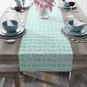 Peut inclure: Une nappe blanche avec un motif turquoise répété de figures et de plantes stylisées. La nappe est posée sur une table en bois dressée pour un repas, avec des assiettes, des bols et des verres visibles.