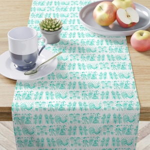 Peut inclure: Chemin de table blanc avec un motif répétitif de personnages et d'éléments floraux turquoise. Le chemin de table est rectangulaire et posé sur une table en bois. Une tasse, une soucoupe, une cuillère et des pommes sont également sur la table.