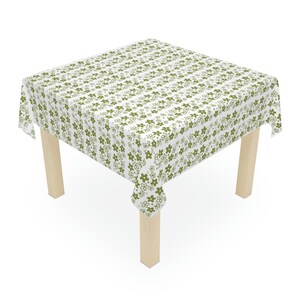 May include: A white tablecloth with a repeating pattern of olive green flowers covers a square table. The table has four light-colored wooden legs. The floral design is evenly spaced across the fabric, creating a cohesive and decorative look.
