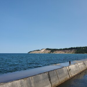 May include: A coastal scene with a concrete pier stretching into a blue sea under a clear sky. A sandy cliff, topped with green trees, is visible in the distance. A person is walking on the pier. The image evokes a sense of tranquility.