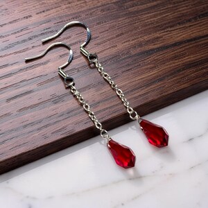 May include: Dangle earrings with silver-tone hooks and chains. Each earring features a faceted, teardrop-shaped, deep red gemstone pendant. The earrings are displayed on a dark wood surface.
