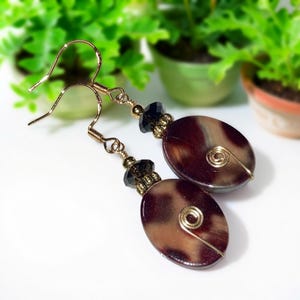 May include: Gold-tone dangle earrings with oval, brown and tan marbled beads. Each earring features a gold wire spiral and black faceted beads. The earrings have gold-colored fish hook ear wires.