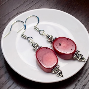 May include: A pair of red oval earrings with silver-colored accents. The earrings have a hook closure and are displayed on a white plate. The earrings are a deep red color and have a smooth, polished surface. The silver accents include decorative beads and ornate end caps.
