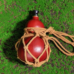 Puede incluir: Una botella de calabaza roja con un tap&oacute;n negro, envuelta en una red gruesa de cuerda marr&oacute;n. La botella est&aacute; sentada sobre un lecho de musgo verde.