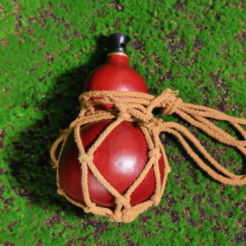 Puede incluir: Una botella de calabaza roja con un tap&oacute;n negro, envuelta en una red gruesa de cuerda marr&oacute;n. La botella est&aacute; sentada sobre un lecho de musgo verde.