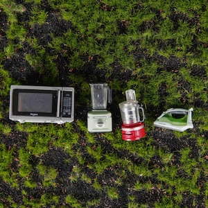 May include: A miniature silver microwave, a light green blender, a red food processor, and a green and white iron. These four small kitchen and household appliances are arranged in a horizontal row on a textured green surface.