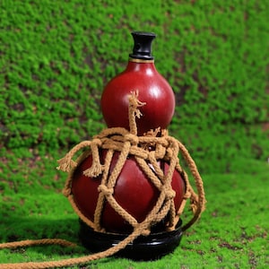 May include: A red gourd bottle with a black stopper, wrapped in a tan rope net. The bottle is sitting on a wooden base.