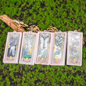 May include: Five pink rectangular keychains with gold-colored chain attachments. Each keychain features a different illustration and text in English and Japanese. The illustrations include a crystal, a bird, a butterfly, a cloud, and gears.