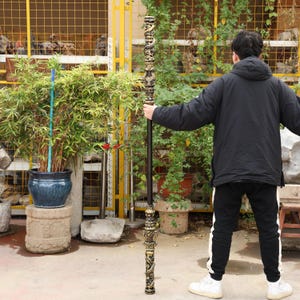 May include: A tall, ornate staff with intricate gold-colored carvings and a dark brown shaft. The staff is held upright, showcasing its detailed design. The carvings appear to be of dragons or mythical creatures. The staff is approximately 6 feet tall.